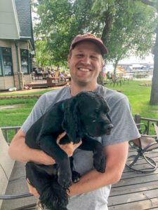 pet-sitting-holding-dog A man is holding a large black puppy with a lake and house in the background.