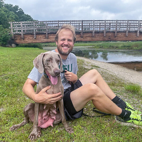 dog-walking-park A man sitting on the ground, arm around a dog, with a bridge in the background.